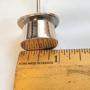 May include: A close-up of a silver metal object with a cylindrical base and a thin, vertical rod. The object is next to a wooden ruler with the text "ACME MADE IN CANADA". The ruler measures approximately 2.5 cm.
