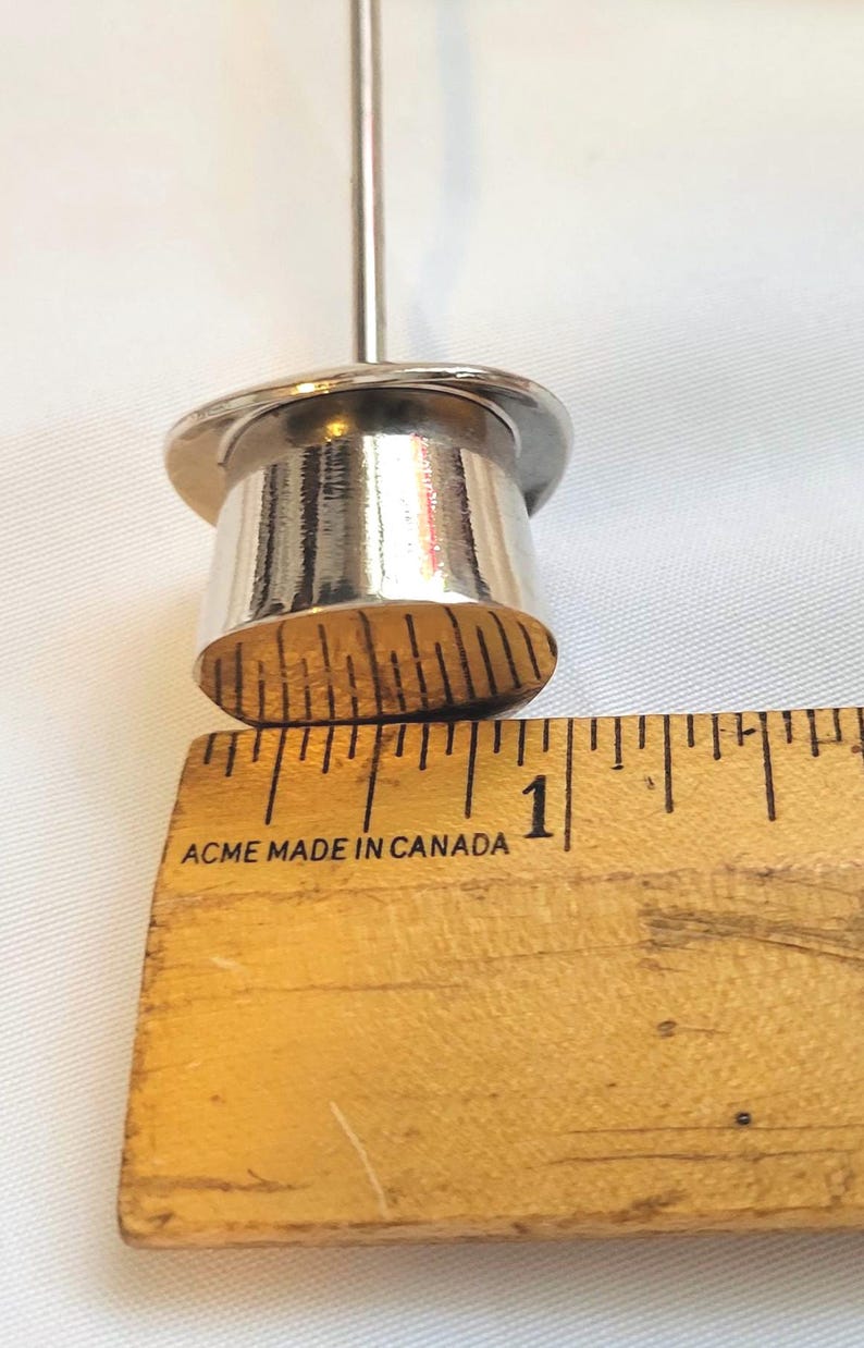 May include: A close-up of a silver metal object with a cylindrical base and a thin, vertical rod. The object is next to a wooden ruler with the text "ACME MADE IN CANADA". The ruler measures approximately 2.5 cm.