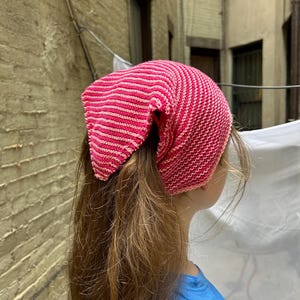 Puede incluir: Una diadema de punto a rayas rosas y blancas. La diadema está envuelta alrededor de la cabeza. La persona lleva una camiseta azul.