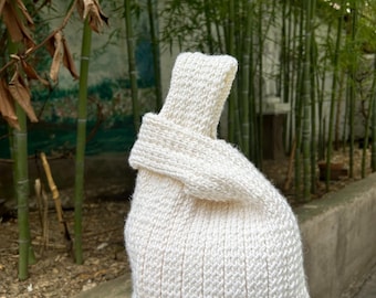 May include: A cream-colored, hand-knitted bag with a knotted handle. The bag is made of thick yarn and has a textured appearance. The bag is sitting on a wooden bench in front of a bamboo background.