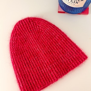 Könnte beinhalten: Eine leuchtend rote Strickmütze. Die Mütze ist aus einem weichen, strukturierten Garn gefertigt und hat ein schlichtes, klassisches Design. Die Mütze wird auf einer weißen Oberfläche gezeigt. Eine Flasche Irish Gin ist im Hintergrund.