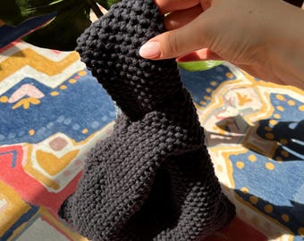May include: A dark gray, hand-knitted headband with a twisted knot design. The headband is made of a thick, textured yarn and is held by a hand. The background includes a colorful patterned fabric and a large green leaf.