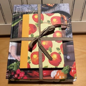May include: A wrapped gift set with a brown ribbon. The gift set includes a cookbook with a colorful cover featuring vegetables and a smaller notebook with a yellow cover and a pattern of red apples.
