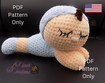 Puede incluir: Un patrón de muñeca de crochet en formato PDF. La muñeca está tumbada de lado con los ojos cerrados. La muñeca está hecha con hilo de color azul y melocotón. El patrón está disponible para descargar.