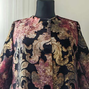Può includere: Una giacca di velluto nero con un motivo floreale in tonalità oro, rosa e marrone. La giacca ha una chiusura con bottoni e un colletto.
