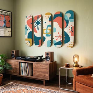 May include: Five skateboard decks mounted on a wall, each featuring a cream background with abstract shapes in teal, coral, and mustard yellow. The designs include geometric patterns, starbursts, and dots. A framed Talking Heads poster is nearby.