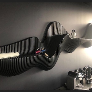 Op de afbeelding: Een lange, zwarte wandplank met een doorlopende, golvende structuur gemaakt van verticale latten. De plank bevat een klein wit astronautenfiguurtje en een meerkleurig handapparaat, met witte rechthoekige objecten aan de linkerkant.