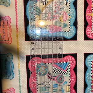 May include: A close-up of a fabric with a repeating pattern of colourful frames with sewing-themed illustrations. The fabric is white with a light grey polka dot pattern. A clear ruler is placed over the fabric, with the numbers 10 to 22 visible.
