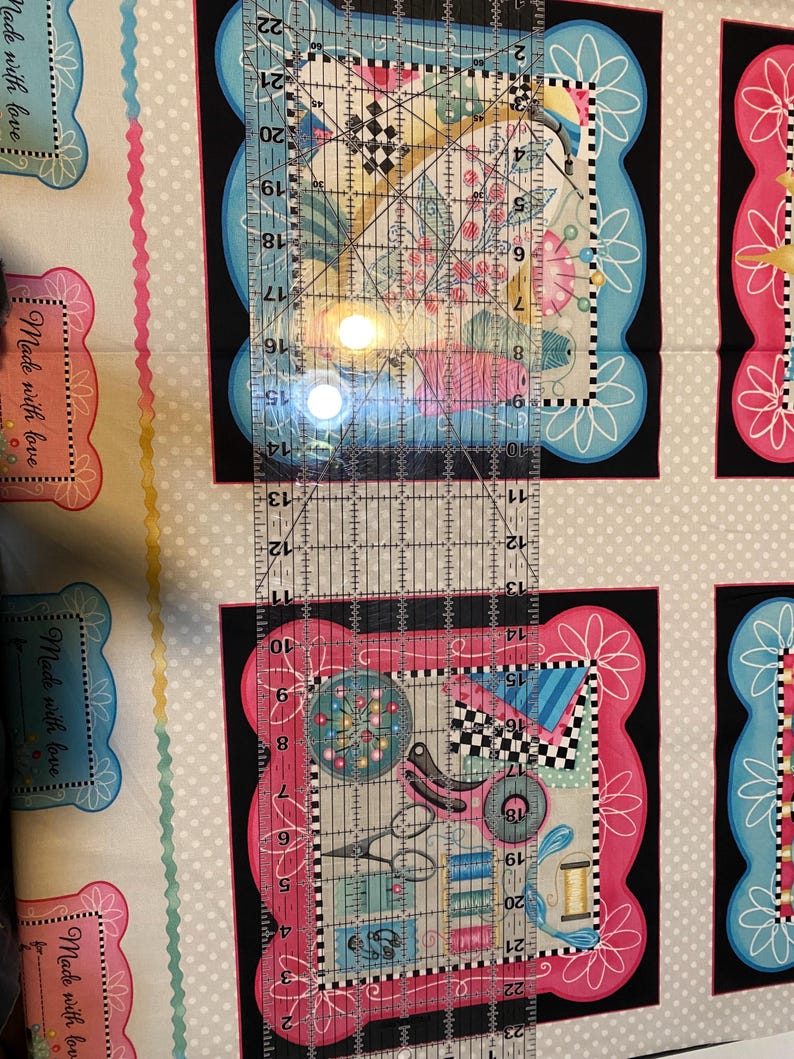 May include: A close-up of a fabric with a repeating pattern of colourful frames with sewing-themed illustrations. The fabric is white with a light grey polka dot pattern. A clear ruler is placed over the fabric, with the numbers 10 to 22 visible.