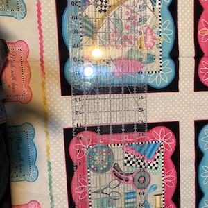 May include: A close-up of a clear ruler with black markings, measuring up to 22 inches. The ruler is placed on top of a fabric with a repeating pattern of pink and blue frames with the text "Made with love" inside each frame.
