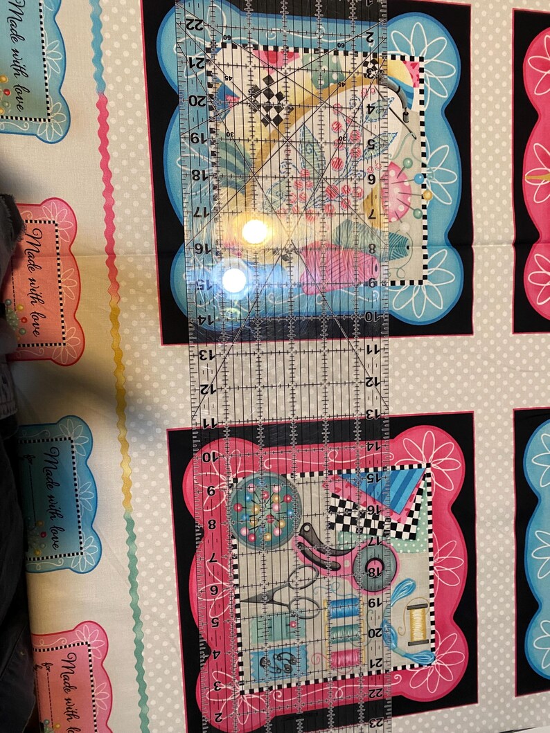May include: A close-up of a clear ruler with black markings, measuring up to 22 inches. The ruler is placed on top of a fabric with a repeating pattern of pink and blue frames with the text "Made with love" inside each frame.