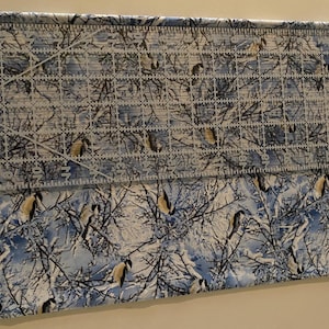 K&ouml;nnte beinhalten: Ein Nahaufnahme eines blauen und wei&szlig;en Stoffes mit einem winterlichen Muster aus B&auml;umen und V&ouml;geln. Der Stoff wird mit einem Lineal gemessen.