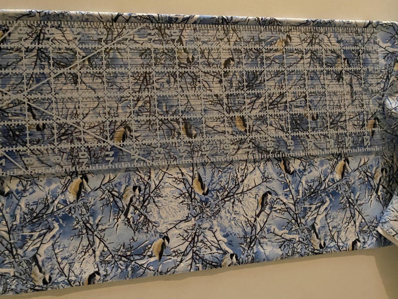 K&ouml;nnte beinhalten: Ein Nahaufnahme eines blauen und wei&szlig;en Stoffes mit einem winterlichen Muster aus B&auml;umen und V&ouml;geln. Der Stoff wird mit einem Lineal gemessen.