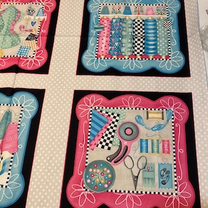 May include: A fabric panel with four squares featuring a sewing theme. Each square has a pink and blue border with a white floral design. The squares depict various sewing supplies, including spools of thread, scissors, and buttons.