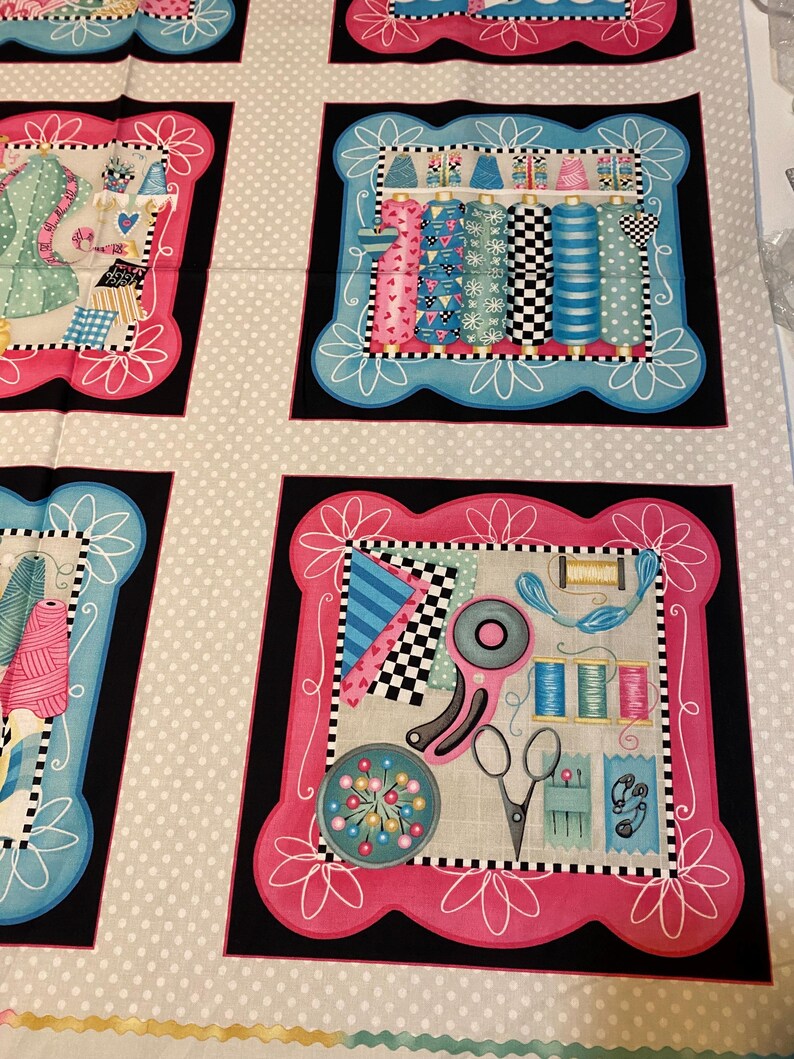 May include: A fabric panel with four squares featuring a sewing theme. Each square has a pink and blue border with a white floral design. The squares depict various sewing supplies, including spools of thread, scissors, and buttons.