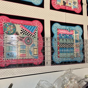 May include: A close-up of a fabric with a repeating pattern of colourful squares and circles. The fabric is framed by a black border and a pink and white border. A ruler is placed on top of the fabric, showing measurements in inches.