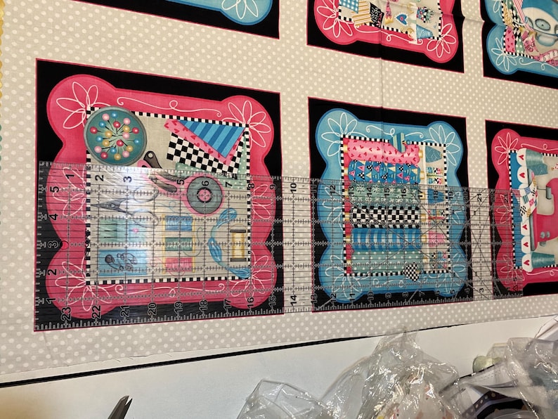 May include: A close-up of a fabric with a repeating pattern of colourful squares and circles. The fabric is framed by a black border and a pink and white border. A ruler is placed on top of the fabric, showing measurements in inches.