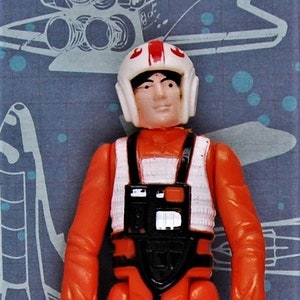 May include: A vintage action figure of a Star Wars pilot wearing an orange jumpsuit with white accents, a black and white flight suit, and a white helmet with a red stripe. The figure has a black belt and a black and white chest piece.