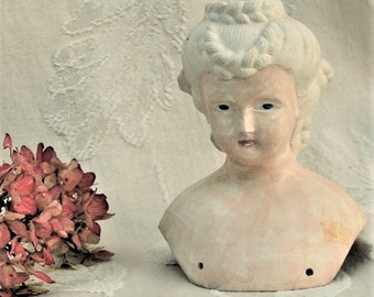 May include: A vintage porcelain doll head with a white painted hairstyle and a pale pink face. The doll head is missing the body and has two small holes on the chest.