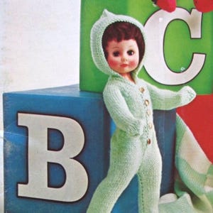 May include: A vintage doll in a light green knitted hooded jumpsuit with wooden buttons. The doll is next to blue and green wooden blocks with the letters "B" and "C". A knitted blanket is partially visible. The jumpsuit is a light green colour.