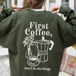 Puede incluir: Sudadera verde militar con el texto blanco "First Coffee, then I do the things". El diseño incluye una cafetera, una taza de café y un jarrón con flores. La sudadera es de corte holgado.