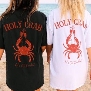 May include: Black and white t-shirts with a red crab graphic, champagne glasses, and the text "HOLY CRAB" and "Let's Get Crackin'!" on the back. The shirts are worn on a beach.
