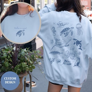May include: Pale blue sweatshirt with a custom design of marine animals in light blue. The front displays a turtle graphic and the words "Kindness is a current". The back features a whale, octopus, and other sea creatures.