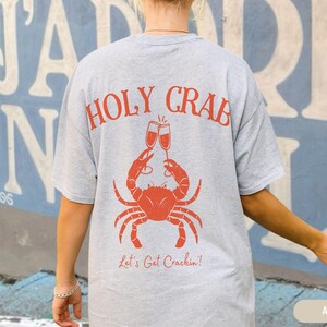 May include: Ash-colored t-shirt with a red crab graphic holding champagne glasses. The text "HOLY CRAB" arches above the crab, with "Let's Get Crackin'!" below. The shirt is a casual, comfortable style.