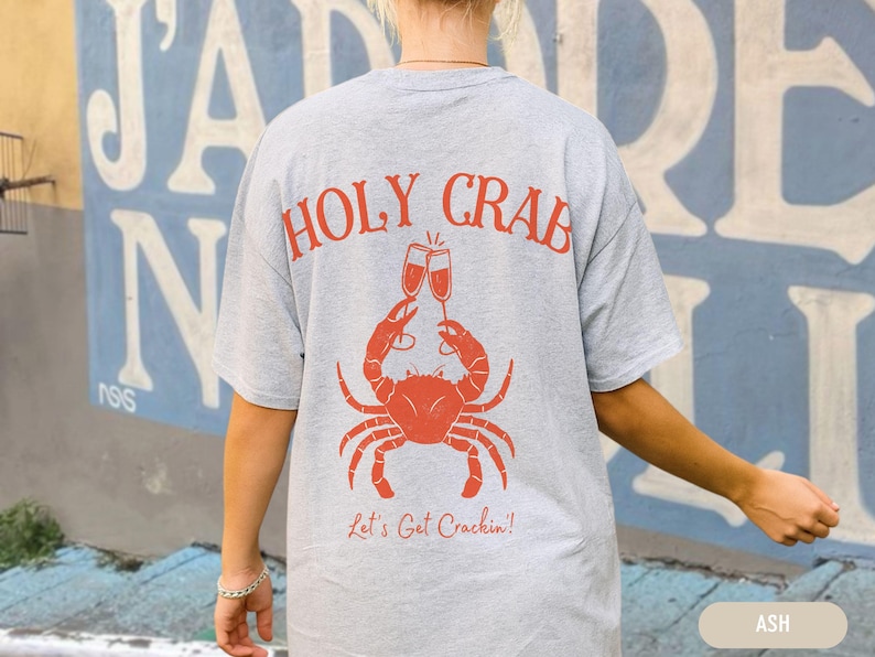 May include: Ash-colored t-shirt with a red crab graphic holding champagne glasses. The text "HOLY CRAB" arches above the crab, with "Let's Get Crackin'!" below. The shirt is a casual, comfortable style.