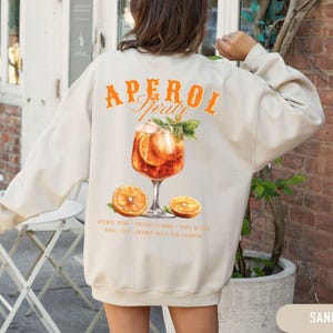 Puede incluir: Sudadera color arena con un gráfico naranja de "Aperol Spritz". El gráfico presenta una ilustración de acuarela del cóctel, rodajas de naranja y texto. La sudadera es de estilo casual y cómodo.