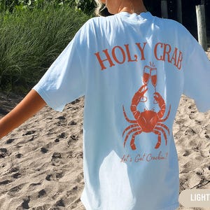 May include: Light blue t-shirt with an orange crab graphic holding champagne glasses. The text "HOLY CRAB" arches above the crab, with "Let's Get Crackin'!" below. The shirt is a relaxed fit.