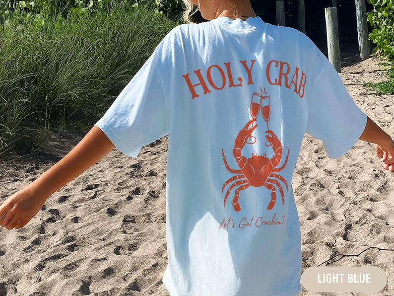 May include: Light blue t-shirt with an orange crab graphic holding champagne glasses. The text "HOLY CRAB" arches above the crab, with "Let's Get Crackin'!" below. The shirt is a relaxed fit.