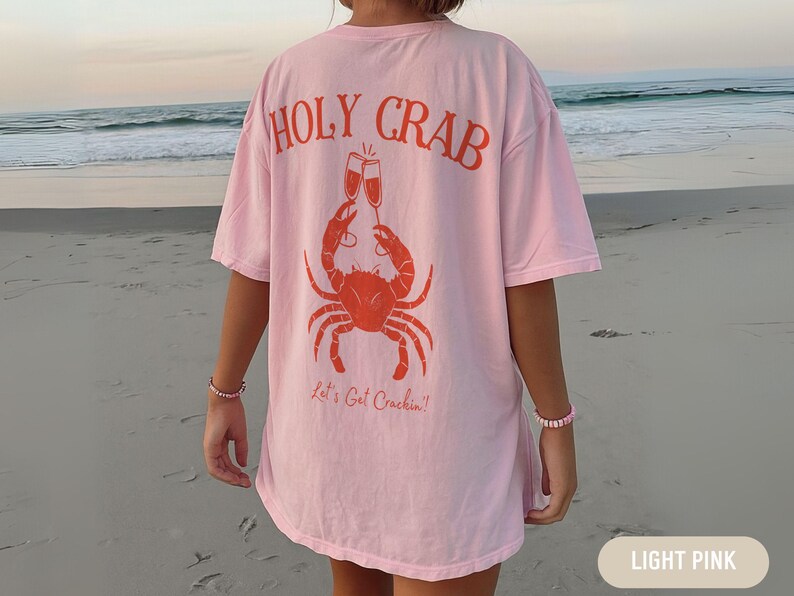 May include: Light pink t-shirt with the words "HOLY CRAB" in orange, above a crab illustration holding two champagne glasses. The text "Let's Get Crackin'!" is below the crab. The shirt is worn on a beach.