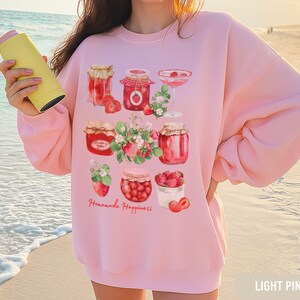 Puede incluir: Sudadera rosa claro con un diseño de acuarela de tarros de mermelada de fresa, fresas y flores. El texto "Homemade Happiness" está escrito en cursiva. La sudadera es de color rosa claro.