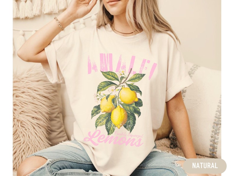 Pu&ograve; includere: Una maglietta bianca con una grafica rosa di limoni e il testo "Avalon Lemons" stampato sul davanti.