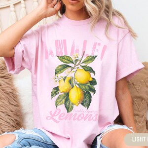 Pu&ograve; includere: Una maglietta rosa chiaro con una grafica di limoni su un ramo. Il testo "Awaken" e "Freshly Squeezed Lemons" &egrave; stampato sulla maglietta.