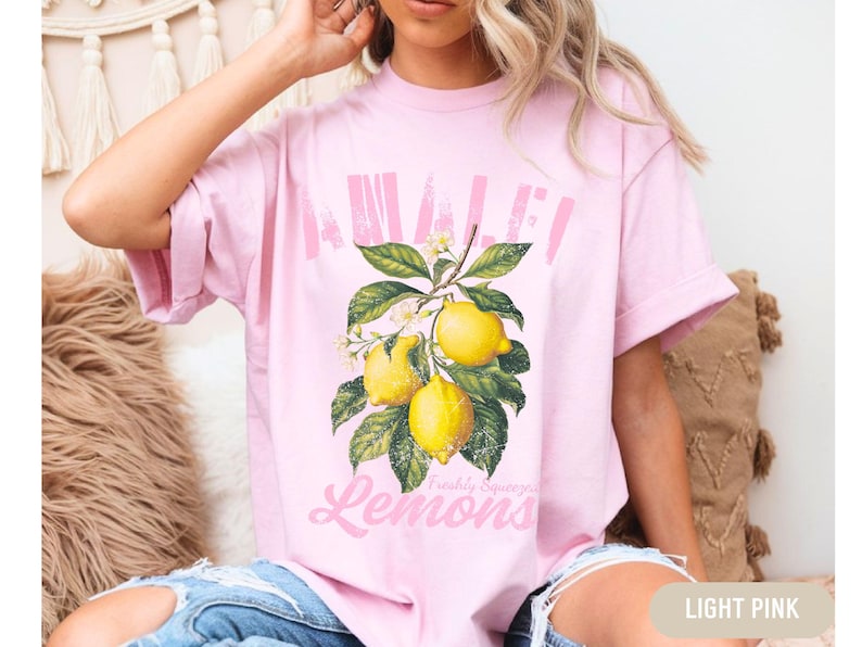 Pu&ograve; includere: Una maglietta rosa chiaro con una grafica di limoni su un ramo. Il testo "Awaken" e "Freshly Squeezed Lemons" &egrave; stampato sulla maglietta.