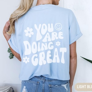 Op de afbeelding: Een lichtblauw T-shirt met een witte afbeelding die "You are doing great" zegt met een smiley en bloemen.
