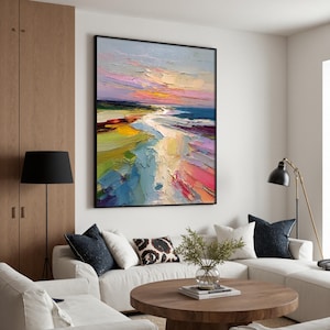 May include: Large framed abstract oil painting of a beach scene. The artwork features a vibrant palette of colors, including blues, pinks, yellows, and greens, depicting a sunset over the ocean. The painting is displayed in a modern living room setting.