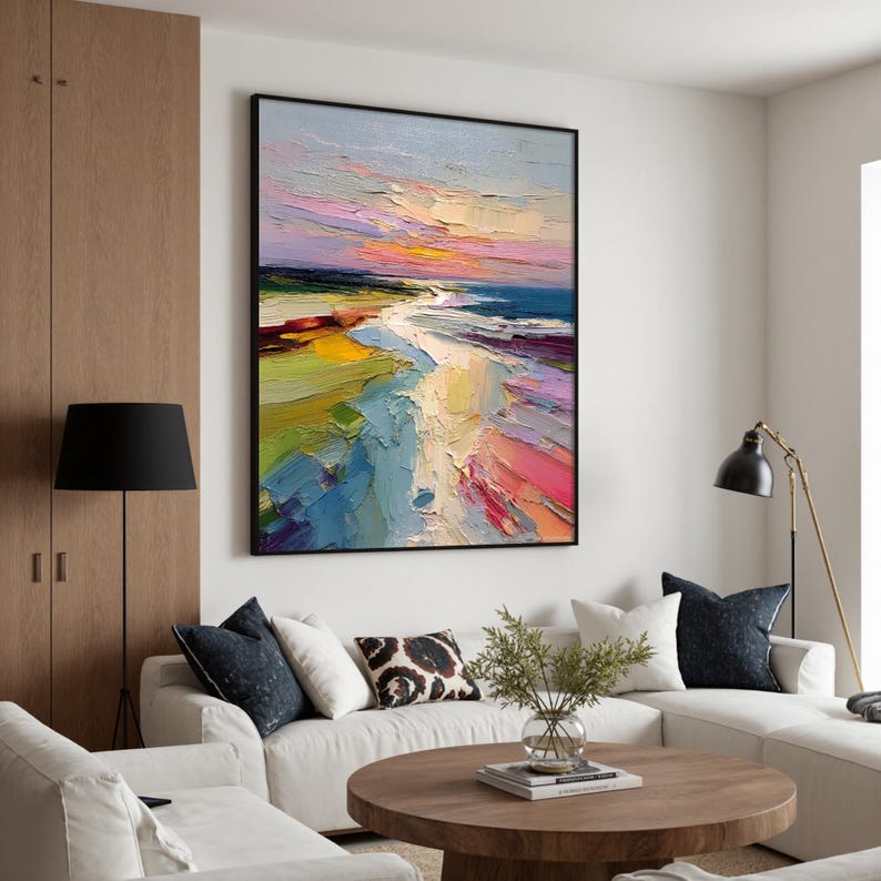 May include: Large framed abstract oil painting of a beach scene. The artwork features a vibrant palette of colors, including blues, pinks, yellows, and greens, depicting a sunset over the ocean. The painting is displayed in a modern living room setting.