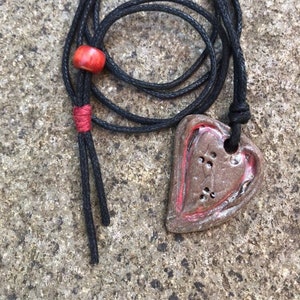 May include: A handmade necklace featuring a heart-shaped pendant with a brown and red glaze. The pendant is suspended from a black cord with a red bead and accents. The necklace is displayed on a textured surface.