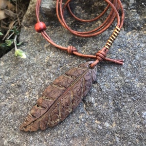 May include: A brown leather necklace with a leaf pendant. The pendant is intricately detailed with a textured surface. The necklace features a red bead and a gold-colored metal accent. The necklace is displayed on a gray stone surface.
