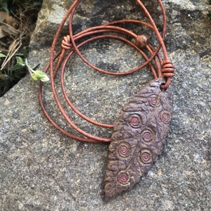 May include: A brown leather necklace with a leaf-shaped pendant. The pendant has a textured surface with spiral designs and red accents. The necklace is displayed on a gray stone surface.