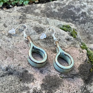 May include: A pair of handmade ceramic earrings. Each earring features a teardrop-shaped design with a light blue glaze and a dark brown rim. The earrings have silver-colored ear wires and clear plastic earring backs.