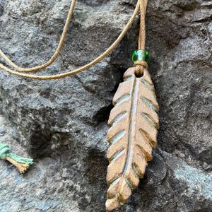 May include: A handcrafted pendant necklace featuring a leaf-shaped charm. The pendant is a light brown color with etched details and a green accent bead. The necklace cord is a natural tan color.
