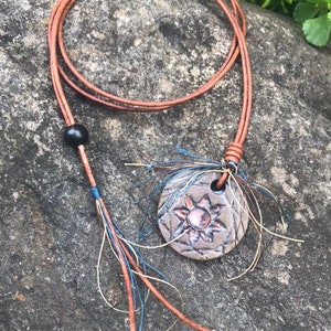 May include: A brown leather necklace with a circular pendant. The pendant is a textured, gray-brown color with a sunburst design in the center. The necklace has a black bead and decorative tassels.
