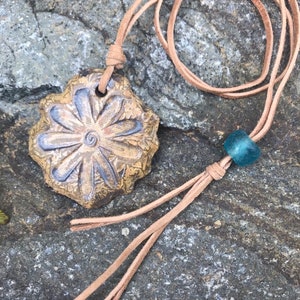 May include: A handcrafted necklace featuring a textured, flower-shaped pendant with blue accents. The pendant hangs from a tan leather cord, with a blue bead and decorative tassels. The necklace is displayed on a stone surface.