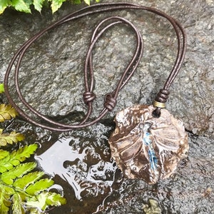 May include: A handcrafted ceramic pendant necklace with a brown leather cord. The pendant is a rough, organic shape with a dragonfly design in blue and brown. The necklace is displayed on a wet, dark stone surface.