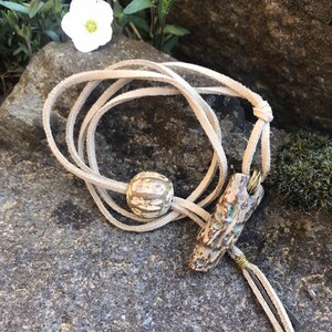 May include: A handmade necklace featuring a cream-colored suede cord. The necklace has a ceramic bead and a ceramic pendant with blue and brown accents. The pendant is attached with gold-colored wire. The necklace is displayed on a gray stone surface.