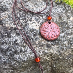 May include: A handmade necklace featuring a round, reddish-pink pendant with a floral design. The pendant is suspended from a brown cord, accented with orange beads. The necklace is displayed against a textured gray stone background.